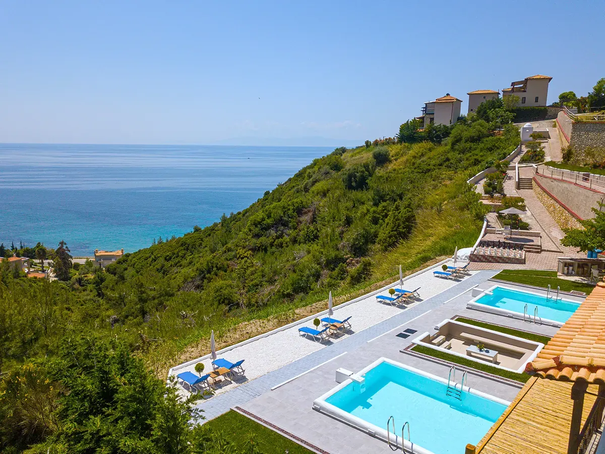 Aerial view of Koumaros ApartHotel with its pools on the green hillside above the sea